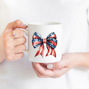 May include: White ceramic mug with a red, white, and blue bow printed on the front. The bow is made of ribbon with stars and stripes.