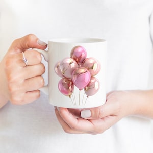 May include: White ceramic mug featuring a cluster of pink and gold metallic balloons. The balloons have a glossy finish and are tied together with gold strings. The mug is held by a person.