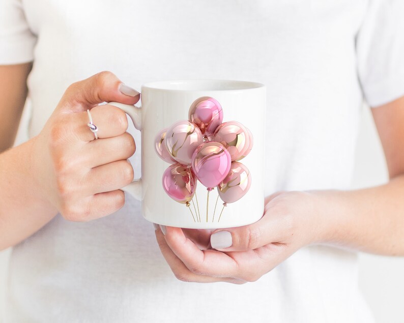 May include: White ceramic mug featuring a cluster of pink and gold metallic balloons. The balloons have a glossy finish and are tied together with gold strings. The mug is held by a person.