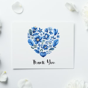 May include: A white greeting card with a blue floral heart design and the words "Thank You" in black script. White flowers and petals surround the card on a white background.