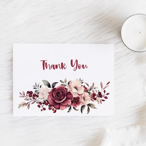 May include: A white card with the words "Thank You" in burgundy script. The card features a watercolor floral design in shades of burgundy, pink, and cream. A lit candle in a glass jar sits to the right.