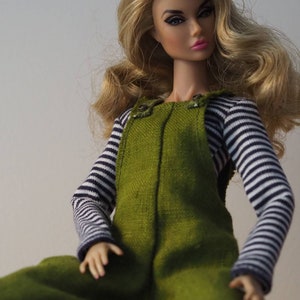 Peut inclure: Une poupée aux longs cheveux blonds portant une salopette verte avec une chemise rayée. La poupée a une lèvre rose et regarde la caméra. La citation "Rien ne peut ternir la lumière qui brille de l'intérieur. -Maya Angelou" est écrite sous la poupée.