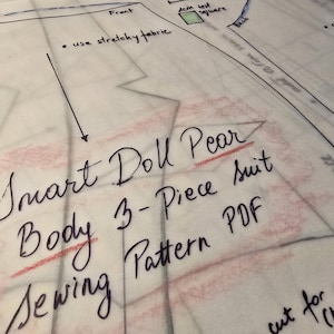 May include: A translucent sewing pattern sheet for a Smart Doll Pear 3-Piece suit. The paper features black handwritten text, including the title, instructions to use stretchy fabric, and a 4cm test square. Lines delineate pattern pieces.