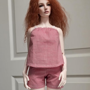 May include: A ball-jointed doll with long, wavy red hair wearing a two-piece outfit consisting of a sleeveless, red and white gingham patterned top with thin white straps and matching red and white gingham shorts with visible white stitching.