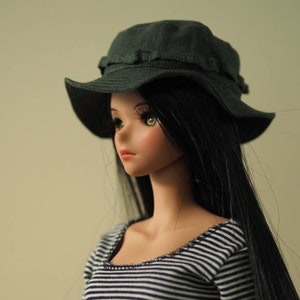 Puede incluir: Una muñeca con el pelo largo y negro que lleva un sombrero de pescador verde oscuro y una camisa a rayas.