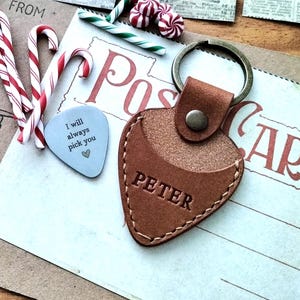 May include: A brown leather guitar pick keychain with the name "PETER" embossed on it, resting on a postcard. Candy canes and a guitar pick with the text "I will always pick you" are also present.