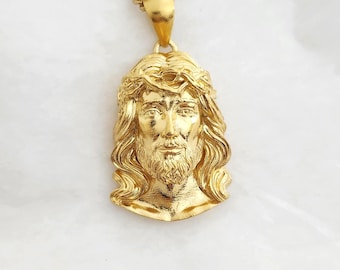 May include: A gold-toned metal pendant necklace featuring a detailed, three-dimensional relief portrait of a figure with long hair and a beard, wearing a crown of thorns. The pendant is suspended from a matching gold-toned metal chain link necklace.