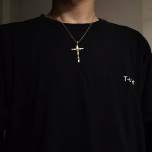 May include: A person wearing a black t-shirt with the word "T-shirt" printed on it. They are wearing a gold chain necklace with a gold crucifix pendant.