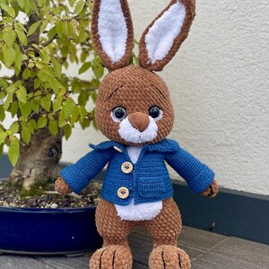 May include: A brown crocheted rabbit wearing a blue crocheted jacket with brown buttons. The rabbit has white fur on its ears and a white patch on its chest.