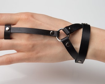 May include: Black leather hand harness with a finger loop and wrist straps. The harness features silver-tone metal hardware, including rivets and a connecting ring. The design is minimalist and modern, suitable for alternative fashion.