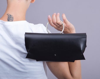 May include: A black leather clutch with a silver ring handle. The clutch has a flap closure and a minimalist design.