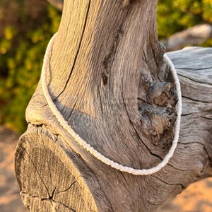 May include: A white beaded necklace made of small, round beads. The necklace is draped over a piece of weathered wood.