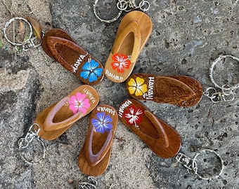 May include: Six wooden flip-flop keychains with silver key rings. Each keychain features a different colored hibiscus flower and the word "Hawaii!" written on the strap.