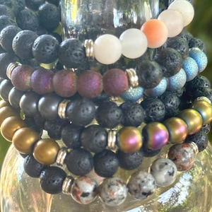 May include: A stack of beaded bracelets in various colors and textures. The bracelets feature beads in black, gold, iridescent, and speckled patterns. Some bracelets have white, pink, and orange beads. The bracelets are held together with silver-colored metal.