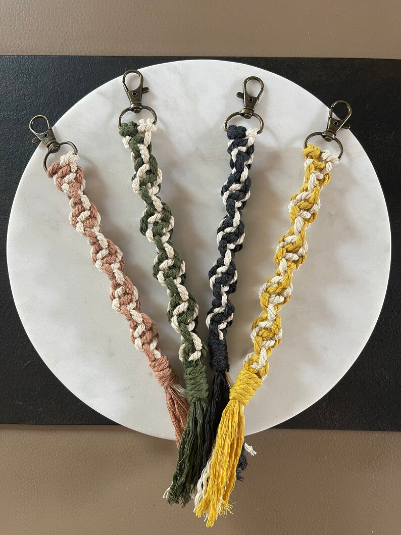 May include: Four handmade macrame keychains with tassels. The keychains are in black, green, yellow, and pink. Each keychain has a white cord woven into the design.