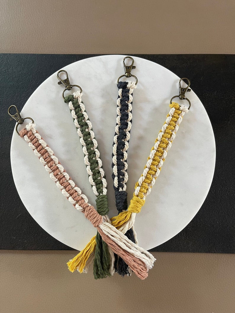 Op de afbeelding: Vier handgemaakte macram&eacute; sleutelhangers met kwastjes in geel, groen, zwart en roze. De sleutelhangers hangen aan een wit marmeren oppervlak.