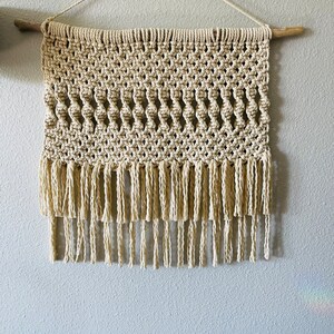 May include: A beige macrame wall hanging with a wood dowel at the top and fringe at the bottom.