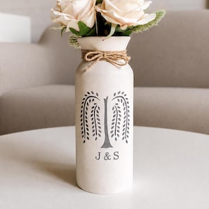 Könnte beinhalten: Eine cremefarbene Keramikvase mit einem grauen Trauerweiden-Design, mit Bindfaden gebunden und gefüllt mit Kunstrosen und getrockneten Gräsern. Die Vase trägt die Initialen "J & S".
