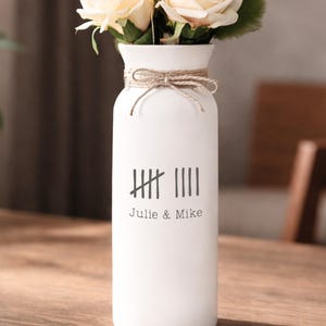 May include: A white ceramic vase with a jute twine accent and a bouquet of cream-colored roses. The vase has a tally mark design and the names "Julie & Mike" printed on it. The vase is on a wooden surface.