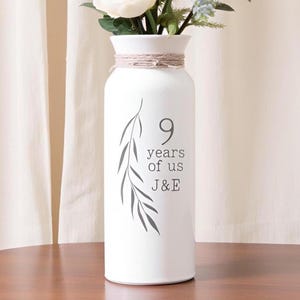 May include: White vase with a floral arrangement. The vase has a cylindrical shape with a narrow neck, wrapped with twine. The text "9 years of us J&E" is printed on the front, along with a leaf design. The vase is on a wooden surface.
