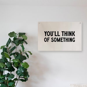 May include: A white canvas wall hanging with black text that reads "You'll think of something". The hanging is displayed on a white wall with a green plant in a black wire basket and a wooden bench with a white cushion.