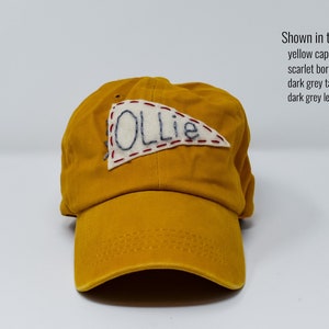 May include: A yellow baseball cap with a white felt pennant sewn on the front. The pennant has the word "OLLIE" embroidered in dark grey thread. The cap has a scarlet border thread, a dark grey tassel thread, and a dark grey letter thread.