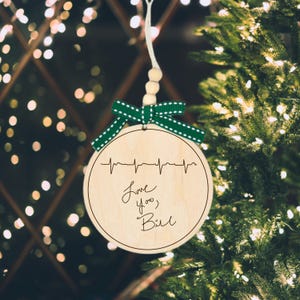 Puede incluir: Un adorno navideño redondo de madera con una cinta verde y blanca punteada y una cuerda de cuentas de madera. El adorno tiene un diseño de línea de latidos del corazón y la frase "Love you, Bill" escrita a mano.