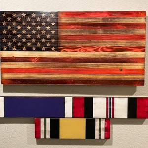 May include: A wooden American flag with red, white, and blue stripes and a field of stars. Below the flag are two wooden military ribbons with red, white, blue, black, and gold stripes.