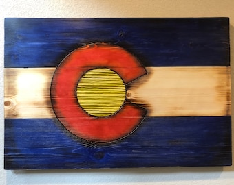 Puede incluir: Arte de pared de madera con la bandera del estado de Colorado. El diseño incluye franjas azules en la parte superior e inferior, una franja blanca central y un emblema rojo y amarillo. La madera tiene un aspecto rústico y envejecido.