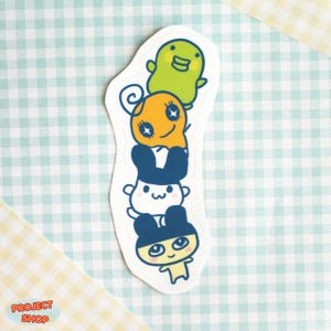 May include: A die-cut sticker featuring a stack of four cartoon characters. The characters are in shades of green, orange, white, and blue. The sticker has a white border and is on a checkered background.