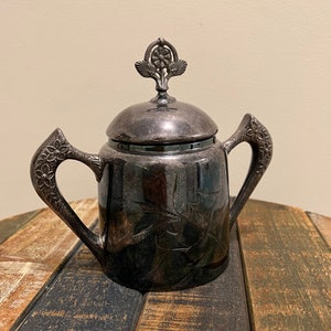May include: A silver sugar bowl with a floral design and a lid with a floral finial. The bowl has two handles and is sitting on a wooden table.