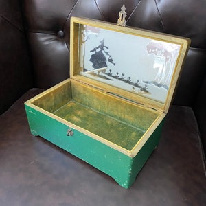 May include: A green wooden box with a hinged lid and a brass handle. The lid is open and reveals a mirror with a silhouette of a woman in a dress and trees.