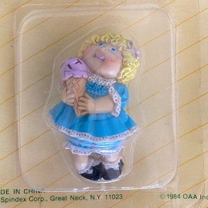 Puede incluir: Una figura de Cabbage Patch Kids en su embalaje original. La figura es de una niña rubia con un vestido azul que sostiene un cono de helado. El embalaje es verde y amarillo con las palabras "FIGURINE" y "Cabbage Patch Kids".