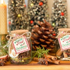 Może przedstawiać: Dwa szklane słoiki z korkowymi wieczkami i sznurkowymi kokardkami, z etykietą "Farm Fresh Christmas Simmering Potpourri". Słoiki wypełnione są przyprawami, suszonymi cytrusami i goździkami. Widoczny jest również duży szyszka i anyż gwiaździsty.