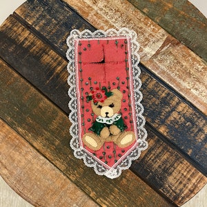 May include: A pink felt bookmark with a beaded border and a felt bear applique. The bear is wearing a green felt dress and has a red felt flower on its head. The bookmark is trimmed with white lace.
