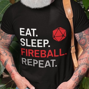 Może przedstawiać: Czarny t-shirt z napisem "EAT. SLEEP. FIREBALL. REPEAT." i czerwoną geometryczną figurą. Koszulka jest noszona przez osobę z tatuażami i brodą.
