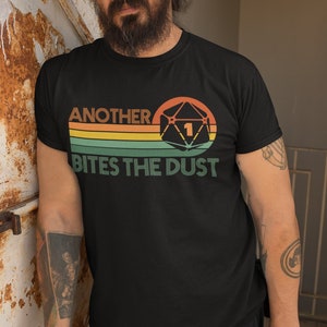 May include: Black t-shirt with a retro-style graphic of a 20-sided die with a 1 showing. The text "Another Bites The Dust" is printed below the die.