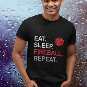 Może przedstawiać: Czarny t-shirt z napisem "EAT. SLEEP. FIREBALL. REPEAT." w kolorze białym i czerwonym oraz czerwonym geometrycznym kształtem. Koszulka jest noszona przez osobę w niebieskich dżinsach.