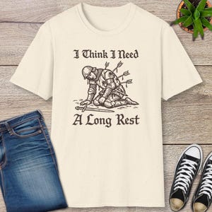 May include: Cream-colored t-shirt featuring a distressed graphic of a medieval knight kneeling, with arrows in his back, and a sword. The text reads "I Think I Need A Long Rest".
