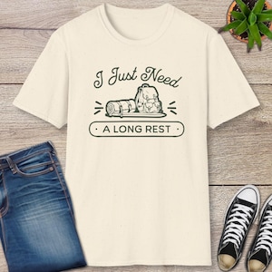 May include: Cream-colored t-shirt with a graphic that reads "I Just Need A Long Rest." The design features a backpack, rolled sleeping bag, and other camping elements. The shirt is paired with blue jeans and black sneakers.
