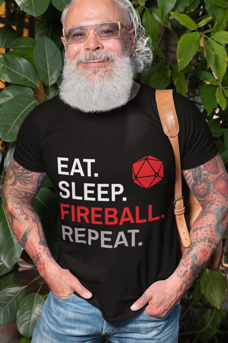 Może przedstawiać: Czarny t-shirt z napisem "EAT. SLEEP. FIREBALL. REPEAT." i czerwoną geometryczną figurą. Koszulka jest noszona przez osobę z tatuażami i brodą.