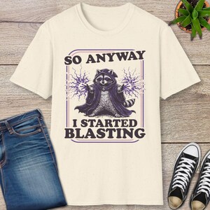 May include: Cream-colored t-shirt featuring a raccoon dressed as a wizard with the text "SO ANYWAY I STARTED BLASTING". The raccoon is wearing a purple robe and conjuring purple lightning bolts. The design is framed in a purple rectangle.