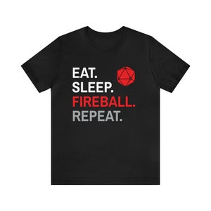 Może przedstawiać: Czarna koszulka z napisem "EAT. SLEEP. FIREBALL. REPEAT." w kolorze białym, czerwonym i szarym. Po prawej stronie tekstu znajduje się czerwona grafika 20-ściennej kostki. Koszulka ma okrągły dekolt i kr&oacute;tkie rękawy.