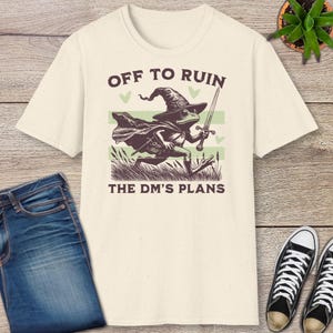 May include: Cream-colored t-shirt featuring a cartoon frog dressed as a wizard, holding a sword, and running. Text reads "OFF TO RUIN THE DM'S PLANS". The design includes a wizard hat, cape, and green accents. The shirt is paired with blue jeans and black sneakers.