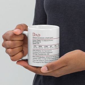 May include: White ceramic mug with a black handle. The mug has a red and black design with the word "DAD" at the top and a character sheet with stats and abilities. The text reads: "DAD, Medium humanoid, chaotic good, Armor Class 13 (Natural Armor), Hit Points 55, Speed 30ft., STR 18 (+4), DEX 12 (+1), CON 14 (+2), INT 14 (+2), WIS 10 (0), CHA 12 (+1), Skills Athletics +6, Survival +2, Condition Immunities frightened, Damage Resistances cold, Aura of Protection. Whenever the Dad or a friendly member within 10 feet of him must make a saving throw, the creature gains a bonus to the saving throw equal to the Dad's Charisma modifier."