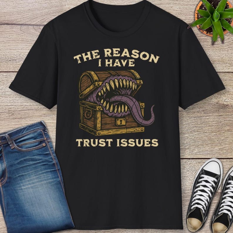 May include: Black t-shirt featuring a graphic of a treasure chest with a monster's mouth and the text "THE REASON I HAVE TRUST ISSUES." The design is in shades of brown, purple, and cream. The shirt is paired with blue jeans and black sneakers.