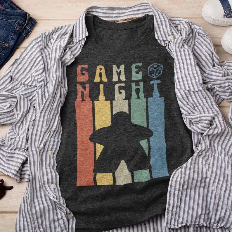 May include: A dark gray t-shirt with a retro-style graphic of a game night icon and the text "GAME NIGHT" in white. The graphic features a rainbow of colors.