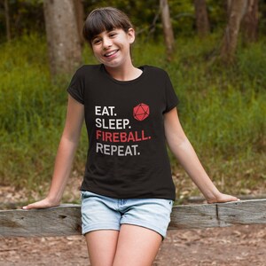 Może przedstawiać: Czarny t-shirt z napisem "EAT. SLEEP. FIREBALL. REPEAT." w kolorze białym i czerwonym. Czerwona grafika wielościanowej kostki do gry znajduje się nad słowem "FIREBALL". Osoba ma na sobie dżinsowe szorty i opiera się o drewniany płot.