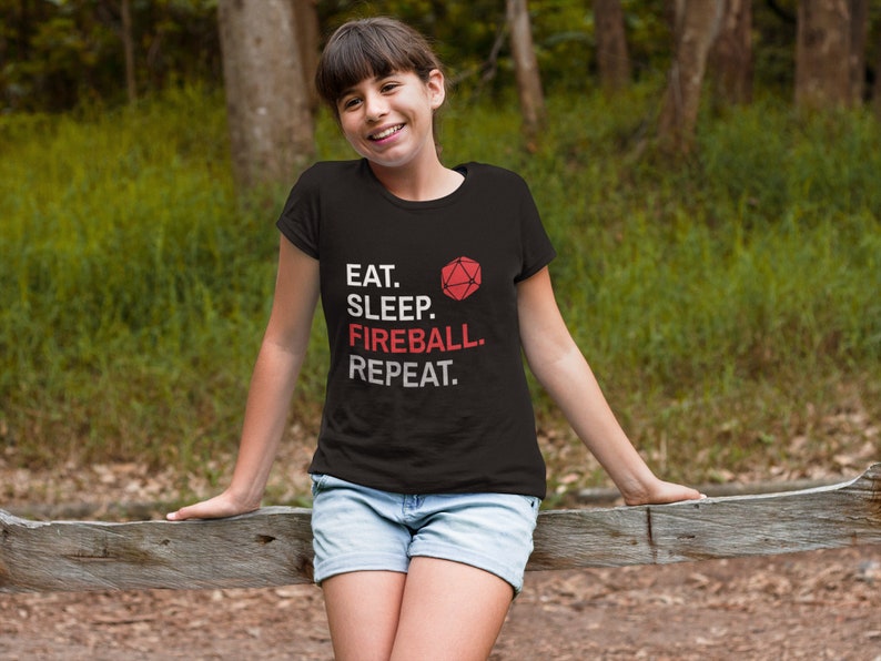 Może przedstawiać: Czarny t-shirt z napisem "EAT. SLEEP. FIREBALL. REPEAT." w kolorze białym i czerwonym. Czerwona grafika wielościanowej kostki do gry znajduje się nad słowem "FIREBALL". Osoba ma na sobie dżinsowe szorty i opiera się o drewniany płot.