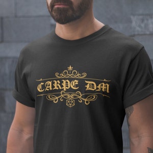 May include: Black t-shirt with gold text and decorative flourishes. The text reads "CARPE DM" with a D20 die graphic. The shirt is a short-sleeved crew neck.